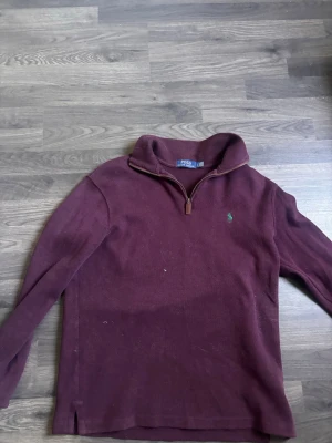 Vinröd halvzip från Polo Ralph Lauren - Vinröd långärmad tröja från Polo Ralph Lauren med halvzip och ståkrage. Tröjan har det klassiska gröna Polo-logot broderat på bröstet och är tillverkad i mjukt material. Perfekt för lager på lager och en chill look.