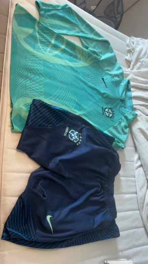 Nike Brasil träningsset blå/turkos - Säljer ett Nike Brasil träningsset med långärmad turkos tröja och mörkblå shorts. Tröjan har grafiskt mönster, halv dragkedja och Brasil-logga. Shortsen är mörkblå med ljusgröna detaljer och elastisk midja. Perfekt för fotbollsträning eller gym.