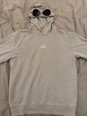 Beige hoodie med glasögon från C.P. Company - Säljer en beige hoodie från C.P. Company med ikoniska inbyggda glasögon i huvan och diskret logga på bröstet. Hoodien har klassisk passform, ribbade muddar och är tillverkad i mjukt bomullsmaterial. Perfekt för dig som gillar streetwear och unika detaljer. Bara att höra av er med prisförslag