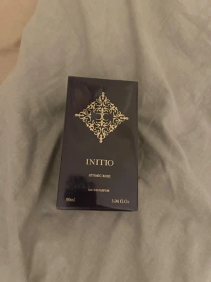 Initio Atomic Rose Eau de Parfum - Initio Atomic Rose Eau de Parfum i en elegant mörklila kartong med gulddetaljer. Flaskan rymmer 90 ml och doften är känd för sina intensiva rosnoter och exklusiva känsla. Förpackningen har en lyxig design med dekorativt mönster framtill.