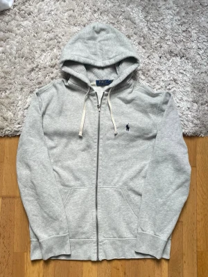 Grå zip hoodie från Polo Ralph Lauren - Snygg grå hoodie med dragkedja från Polo Ralph Lauren. Klassisk design med huva och vita dragsnören, samt den ikoniska mörkblå loggan broderad på bröstet. Tillverkad i mjukt sweatshirtmaterial, perfekt för en chill och avslappnad stil.