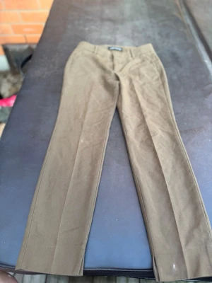 Bruna kostymbyxor från Zara Woman - Snygga bruna kostymbyxor från Zara Woman med klassisk rak passform och pressveck. Byxorna har medelhög midja och är tillverkade i ett mjukt tyg som ger en stilren look. Perfekta för dig som vill ha en clean och tidlös stil.