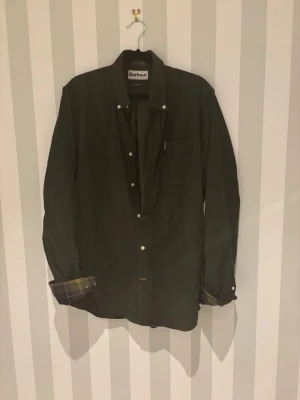 Mörkgrön skjorta från Barbour - Säljer en mörkgrön skjorta från Barbour med klassisk krage och knappar framtill. Skjortan har lång ärm och rutigt foder i ärmsluten för en snygg detalj. Passformen är tailored fit vilket ger en stilren siluett. Storlek M