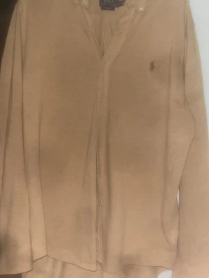 Beige skjorta från Polo Ralph Lauren - Klassisk beige skjorta från Polo Ralph Lauren med knappkrage och broderad logga på bröstet. Skjortan har långa ärmar och knappar framtill. Perfekt för en stilren och avslappnad look.