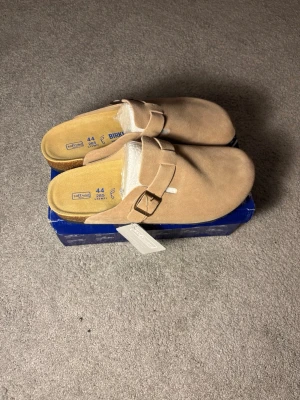 Beige Birkenstock tofflor mocka - Säljer ett par snygga beige tofflor från Birkenstock i mjuk mocka. Modellen har en justerbar rem med metallspänne och en bekväm innersula. Perfekta för chill dagar hemma eller när du vill ha det extra bekvämt.
