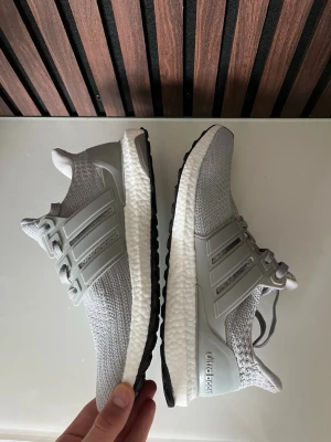 Grå Adidas Ultraboost sneakers - Säljer ett par grå Adidas Ultraboost sneakers med stickad ovandel och klassiska tre ränder på sidan. Skorna har vit Boost-mellansula och svart yttersula. Perfekta för dig som gillar stilrena och bekväma sneakers med modern design.