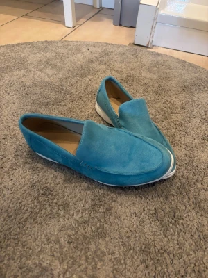 Turkosa loafers i mocka - Säljer ett par stilrena turkosa loafers i mocka med vit sula. Skorna har en klassisk slip-on design och är perfekta för dig som vill ha en färgklick till outfiten. Insidan är beige och sulan är platt för en avslappnad look.