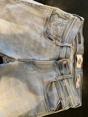 Ljusblå raka jeans från Replay - Säljer ett par ljusblå jeans från Replay med klassisk femficksmodell och raka ben. Jeansen har snygga detaljer som Replay-lapp bak i midjan och broderade bakfickor. Tillverkade i mjuk denim med bälteshällor och knappgylf.