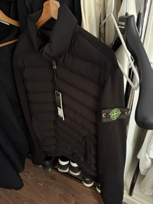 Svart pufferjacka från Stone Island - Säljer en svart pufferjacka från Stone Island med ikonisk patch på vänster ärm. Jackan har hög krage, dragkedja framtill och två sidofickor. Materialet är quiltat och ger en riktigt clean och modern look. Perfekt för kalla dagar.