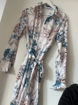 Blommig klänning med knytband - Snygg klänning i vitt med blommigt mönster i blått och brunt. Långärmad modell med krage och knytband i midjan. Perfekt för att chilla hemma i och till fest. Och ger en fräsch och stilren känsla. Tunn och mjuk i materialet, vilket gör den skön att bära.