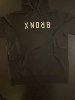 Svart hoodie med BRONX-tryck - Snygg svart hoodie med vit text 'BRONX' på bröstet. Klassisk modell med huva och dragsnören. Perfekt för en avslappnad streetwear-look. Mjuk och bekväm, passar till det mesta i garderoben.