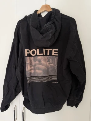 Svart oversized hoodie från Young Poets - Svart hoodie med stor huva och trycket 'POLITE' i beige på ryggen, samt en grafisk bild och text under. Nedtill står det 'YOUNG POETS'. Hoodien är oversized och storlek S.