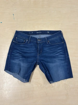 Levis shorts - Säljer ett par klassiska blå levis jeansshorts  med fem fickor och rå, avklippt kant. Shortsen har normal passform och är tillverkade i bomull. Perfekta för sommaren och ger en avslappnad look.