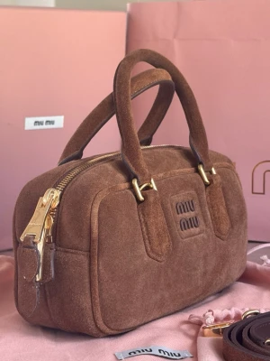 Brun mockaväska från Miu Miu - Snygg handväska i brun mocka från Miu Miu med guldfärgade metalldetaljer och dubbla handtag. Väskan har en klassisk boxig form, dragkedja upptill och diskret logga framtill. Avtagbar axelrem medföljer för extra flexibilitet.