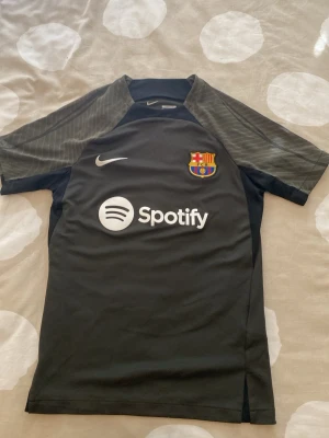 Svart FC Barcelona Nike träningströja - Säljer en svart FC Barcelona träningströja från Nike med klubbmärke och Spotify-logga på bröstet. Tröjan har korta ärmar med gråa detaljer och är tillverkad i ett lätt, andningsbart material som passar perfekt till fotbollsträning.