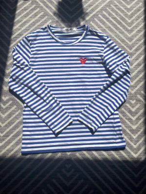 Blåvit randig CDG tröja  - Blå och vit randig långärmad tröja från CDG med rund hals och ett rött broderat hjärta på bröstet. Tröjan är i mjuk bomull och har en avslappnad passform som passar perfekt till jeans eller kjol. Enkel men ändå lekfull design. Myvket eftertraktad och populär tröja!! Det är storlek L men sitter mer som M