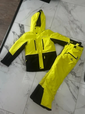 Gula skidbyxor med svart detalj - Snygga skidbyxor och skidjacka från McKinley i starkt gul färg med svarta detaljer vid benslut och fickor. Byxorna har dragkedja och justerbar midja. Perfekta för vinterns alla äventyr i backen eller snön. Både jacka o byxor är i jättefint skick!
