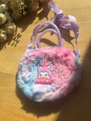 Pastellfärgad fluffig handväska - Söt rund handväska i pastellfärgad fuskpäls med lila, rosa och blå toner. Väskan har två handtag och ett långt lila band för axeln. Framtill sitter en dekoration med figuren Kuromi och texten 'KUNOMI'. Perfekt för dig som gillar kawaii-stil.