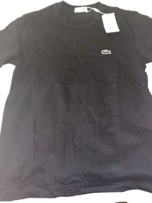 Svart t-shirt från Lacoste - Klassisk svart t-shirt från Lacoste med rund hals och den ikoniska krokodilloggan broderad på bröstet. Regular fit och tillverkad i mjuk bomull som känns skön mot huden. Perfekt basplagg för en clean och stilren look.
