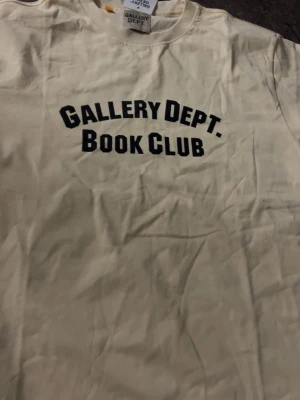 Vit t-shirt Gallery Dept. Book Club - Snygg vit t-shirt från Gallery Dept. med svart text 'Gallery Dept. Book Club' tryckt på bröstet. Klassisk passform och rund halsringning. Perfekt för dig som gillar streetwear och vill ha en enkel men stilren look.