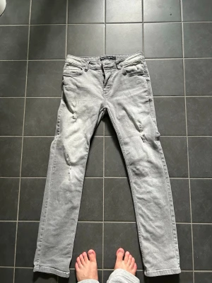 Ljusgrå raka jeans med slitningar - Säljer ett par ljusgrå jeans med raka ben och slitna detaljer på framsidan. Jeansen har klassisk femficksdesign och är tillverkade i bomullsmix för en bekväm känsla. Perfekta för en avslappnad och trendig look.