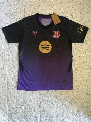 FC Barcelona Bortatröja 26/27 - Snygg FC Barcelona bortatröja i svart och lila med guldfärgade detaljer och Spotifyloggan, klubbloggan och nike logan på framsidan. Perfekt för fotboll eller träning.