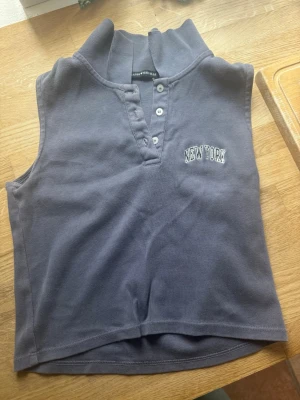 Blå/grå  topp Brandy Melville - Säljer en mörk blå/lite grå ärmlös topp från Brandy Melville med krage och knappar framtill. På bröstet står det 'New York' broderat i vitt. Perfekt till jeans eller kjol för en chill look🩷 Säljer pgr av att den är för liten. Den är i väldigt fint skick men har tvättats någon gång och därmed förlorat lite färg. 