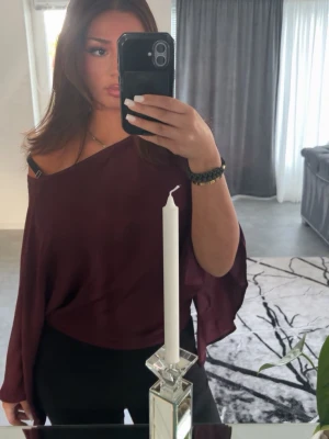 Vinröd offshoulder blus Nelly  - Säljer en vinröd offshoulder blus med lös passform och långa ärmar. Tyget är tunt och faller snyggt över axeln för en avslappnad men trendig look. Perfekt att styla med både jeans och kjol.