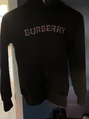 Svart hoodie från Burberry - Snygg svart hoodie från Burberry med klassisk logga broderad i vitt och rött på bröstet. Hoodien har huva och ribbade muddar vid ärmslut och nederkant. Perfekt för dig som vill ha en stilren och exklusiv look.