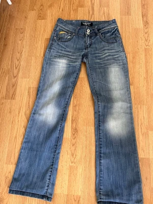 Blå bootcut jeans med slitningar - Snygga blå bootcut jeans med slitningar och ljusa partier på låren och knäna. Jeansen har klassiska fem fickor, dubbla knappar i midjan och kontrastsömmar. Perfekta för en avslappnad och trendig look.