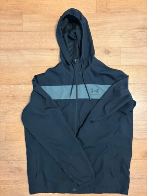 Svart vindjacka från Under Armour - Snygg svart vindjacka från Under Armour med huva och hel dragkedja framtill. Jackan har en grå rand över bröstet och Under Armour-logga på vänster sida. Två sidofickor med dragkedja och elastiska muddar vid ärmsluten. Perfekt för blåsiga dagar.