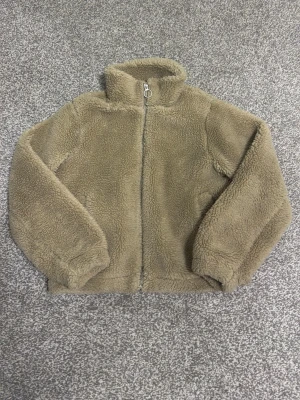 Beige teddyjacka med dragkedja - Mysig beige teddyjacka med hög krage och dragkedja framtill. Jackan har en fluffig och mjuk yta som ger en avslappnad look. Perfekt för kyliga dagar när du vill vara både varm och trendig.