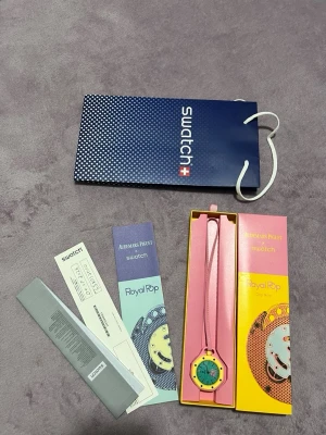Ap X Swatch Rosa Klocka - Unik Swatch Royal Pop klocka med färgglad design. Boetten är rosa med gul ram och urtavlan är turkos med ett coolt mönster. Klockan har ett rosa band och levereras i originalförpackning med tillbehör. Perfekt för dig som gillar statement-accessoarer.