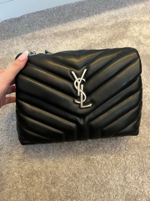 Svart YSL quiltad handväska - Snygg svart quiltad handväska från YSL med silverfärgad kedjerem och ikonisk YSL-logga framtill. Väskan har en mjuk, puffig struktur och elegant chevronmönster. Perfekt accessoar för att lyfta din outfit.