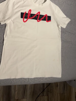 Vit t-shirt med tryck framtill - Vit t-shirt i bomull med korta ärmar och ett svart tryck med röd grafisk detalj på bröstet. Enkel och stilren modell som passar till det mesta. Rund halsringning och normal passform.