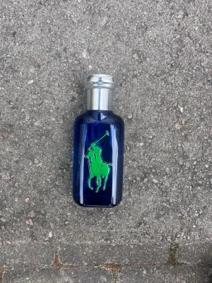Blå parfymflaska med silverlock - Snygg parfymflaska i mörkblått glas med silverfärgat lock. På framsidan finns en grön polospelare och på baksidan en stor gul etta. Flaskan har en modern och sportig design som sticker ut i badrumshyllan.