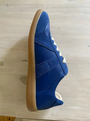 Blå sneakers med mockadetaljer - Snygga blå sneakers med ovandel i både mocka och slätt läder, vita skosnören och klassisk gummisula i brunt. Skon har en låg profil och stilrena linjer som passar till många olika outfits. Det är tyvärr för små för mig så dom är helt oanvända.