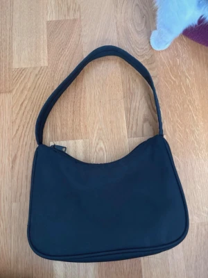 Svart axelväska  - En stilren svart axelväska i klassisk baguette-modell. Väskan har en enkel design med mjukt tyg, dragkedja upptill och bred axelrem. Perfekt för dig som gillar minimalistisk och tidlös stil. Märket är okänt. 