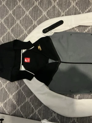 Nike hoodie svart/grå/vit - Snygg hoodie från Nike i svart, grått och vitt med dragkedja framtill. Huva och Nike-logga i guld på bröstet. Ärmarna är vita med en svart ficka på ena armen. Perfekt för en sportig och avslappnad stil.