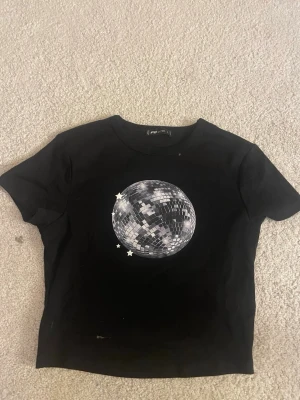 Svart baby tee med discokula  - Cool svart cropped t-shirt från FB Sister med ett stort tryck av en discokula och små stjärnor på bröstet. T-shirten har korta ärmar och är gjord i mjuk bomull, perfekt för en avslappnad och trendig look. Det står att storleken är L men skulle nog säga xs