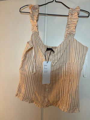 Randig linnetopp från Gina Tricot - Säljer en söt randig linnetopp från Gina Tricot i beige och vit med breda volangband och knappar framtill. Toppen har en avslappnad passform och är perfekt för varma dagar. Materialet känns lätt och luftigt.