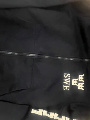 Svart hoodie med SWE-tryck Rosetti - Svart hoodie från Rosetti med dragkedja framtill och vitt SWE-tryck samt tre kronor på bröstet. Hoodien har långa ärmar och vit text på ena ärmen. Perfekt för dig som vill ha en stilren och sportig look.
