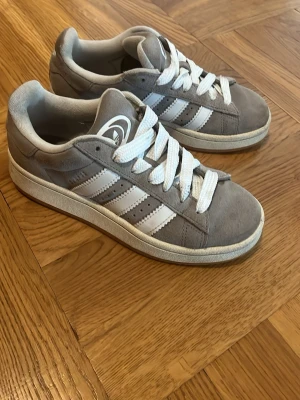 Grå Adidas Campus, storlek 36 - Snygga grå sneakers med klassiska vita ränder på sidorna och vita skosnören. Skorna har en låg profil och en vit sula med brun undersida. Tillverkade i mockaliknande material för en stilren look. Använda men i fint skick som på bilden!