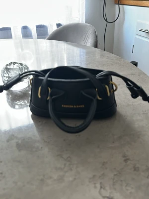 Svart minihandväska med gulddetaljer - Snygg svart minihandväska med guldfärgade metalldetaljer och dubbla handtag. Väskan har en dragskoöppning och stilren design. Perfekt accessoar för dig som gillar trendiga och kompakta väskor.