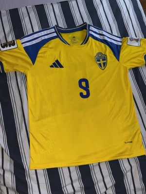 Svensk landslagströja Adidas nr 9 - En helt ny gul och blå svensk landslagströja från Adidas med nummer 9 och namnet ISAK på ryggen. Tröjan har korta ärmar, blå detaljer på axlarna och broderad landslagssköld på bröstet. perfekt för fotbollsträning eller match.