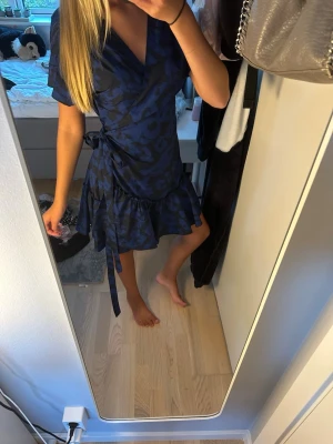 Marinblå omlottklänning från Svea - Snygg marinblå omlottklänning från Svea i storlek XS. Klänningen har knyt i midjan, volangkant nertill och ett diskret mönster i mörkblått. Perfekt för dig som gillar en feminin och trendig stil.
