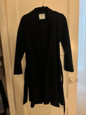 Svart lång kappa med skärp - Stilren svart lång kappa med klassisk krage och tillhörande skärp i midjan. Kappan har raka ärmar och en enkel, tidlös design som passar till många olika outfits. Perfekt för dig som gillar minimalistisk och clean stil.