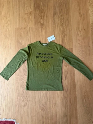 Grön långärmad topp Acne Studios - Snygg grön långärmad topp från Acne Studios med tryck 'STOCKHOLM 1996' på bröstet. Tillverkad i mjuk bomull och har en klassisk, normal passform. Perfekt att styla med jeans eller kjol för en clean look.