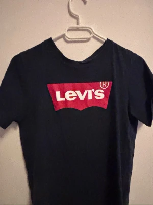Svart Levi's t-shirt med rött tryck - Snygg svart t-shirt från Levi's med det klassiska röda logotrycket på bröstet. T-shirten har rund hals och korta ärmar, perfekt för en avslappnad och trendig stil. Tillverkad i mjuk bomull för skön komfort hela dagen.
