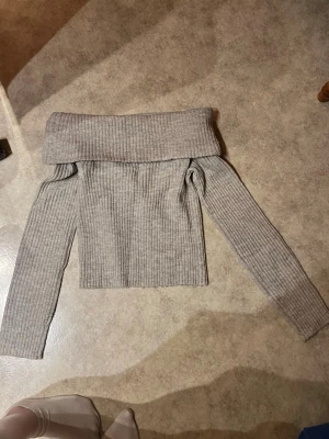 Grå off shoulder/folded stickad tröja - Trendig grå ribbstickad tröja med off shoulder-design och långa ärmar. Tröjan har en bred, vikt krage som ger en snygg och avslappnad look. Perfekt för dig som gillar stilrena och bekväma plagg i garderoben. Använd max en gång säljer då den inte kommer till användning.